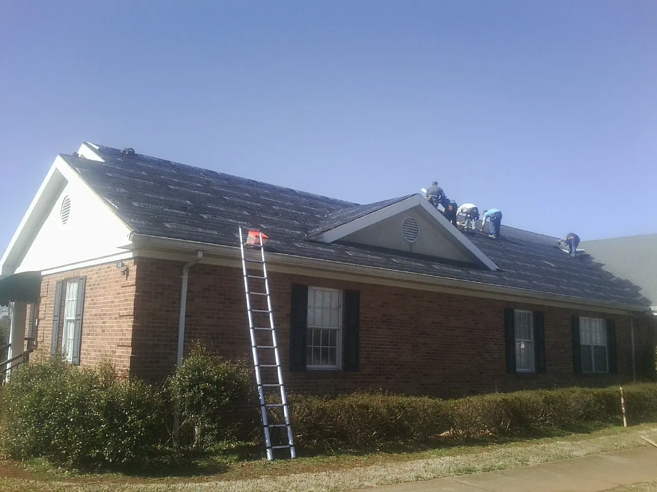 Crew tearing shingles off a brick ranch roof with a ladder set — Metal Roof Repair work by our crew in Girard